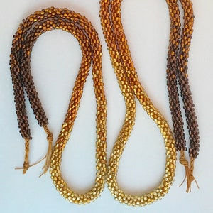 May include: Two beaded necklaces in a gradient of brown to gold. The necklaces are constructed from small, square beads, giving a rope-like texture. Tassels are attached to the ends. The necklaces are displayed against a white background.