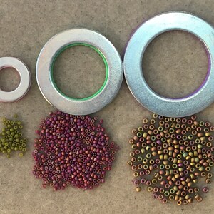 Puede incluir: Tres anillos met&aacute;licos plateados de diferentes tama&ntilde;os, con detalles verdes y morados, se muestran junto a tres montones de peque&ntilde;as cuentas en tonos verdes, morados y dorados. Las cuentas son probablemente para hacer joyas o manualidades.