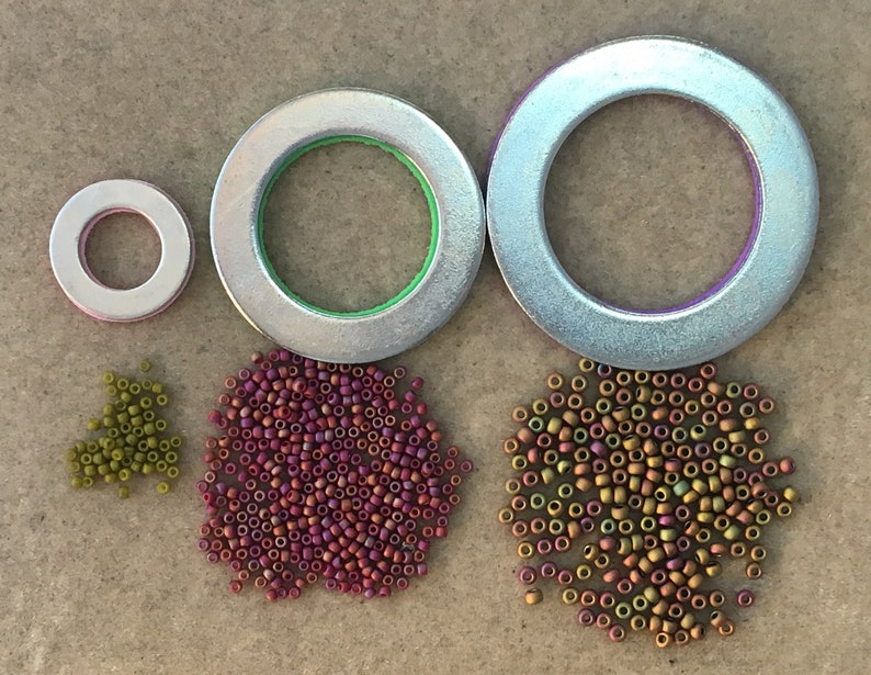 Puede incluir: Tres anillos met&aacute;licos plateados de diferentes tama&ntilde;os, con detalles verdes y morados, se muestran junto a tres montones de peque&ntilde;as cuentas en tonos verdes, morados y dorados. Las cuentas son probablemente para hacer joyas o manualidades.