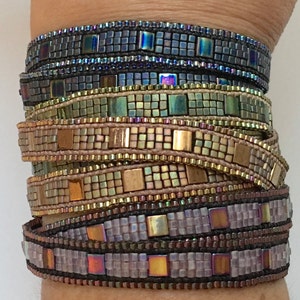 May include: Close-up of several beaded wrap bracelets in various colours. The bracelets are stacked on top of each other, showcasing a variety of iridescent and metallic beads. The colours include shades of grey, green, gold, and purple, with a woven design.