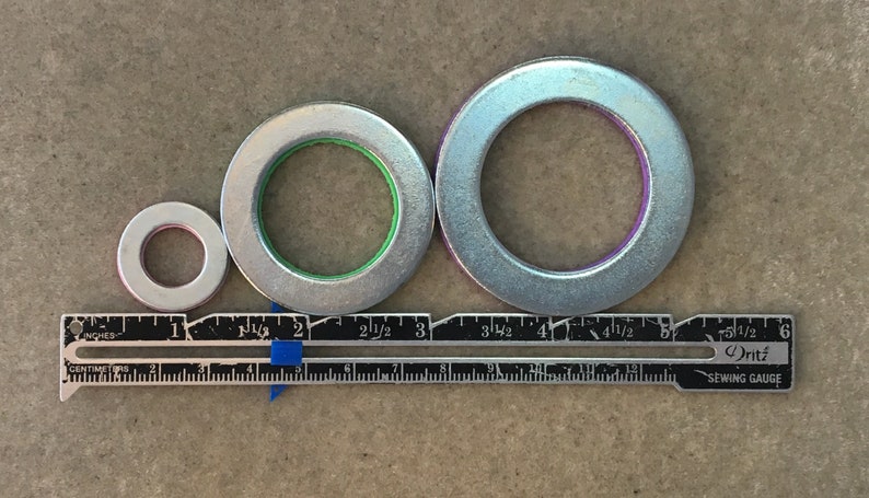 Puede incluir: Tres arandelas met&aacute;licas plateadas de diferentes tama&ntilde;os, con anillos interiores de color rojo, verde y morado. Una regla con medidas en pulgadas y cent&iacute;metros se coloca debajo. La regla est&aacute; marcada con el texto "Dritz Sewing Gauge".