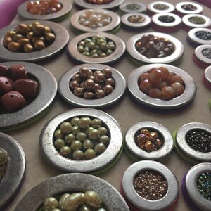 Puede incluir: Una colecci&oacute;n de diversas cuentas y piedras preciosas en peque&ntilde;os recipientes met&aacute;licos redondos. Las cuentas son en tonos marr&oacute;n, dorado, verde y naranja. Los recipientes est&aacute;n dispuestos sobre una superficie clara, con un borde rosa.