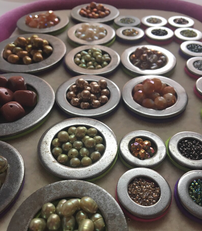 Puede incluir: Una colecci&oacute;n de diversas cuentas y piedras preciosas en peque&ntilde;os recipientes met&aacute;licos redondos. Las cuentas son en tonos marr&oacute;n, dorado, verde y naranja. Los recipientes est&aacute;n dispuestos sobre una superficie clara, con un borde rosa.