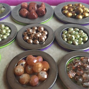 Puede incluir: Una variedad de cuentas de varios colores y formas, exhibidas en peque&ntilde;os recipientes met&aacute;licos redondos. Las cuentas incluyen tonos dorados, marrones, verdes y naranjas, algunas con apariencia facetada o perlada. Los recipientes est&aacute;n sobre fieltro morado y verde.