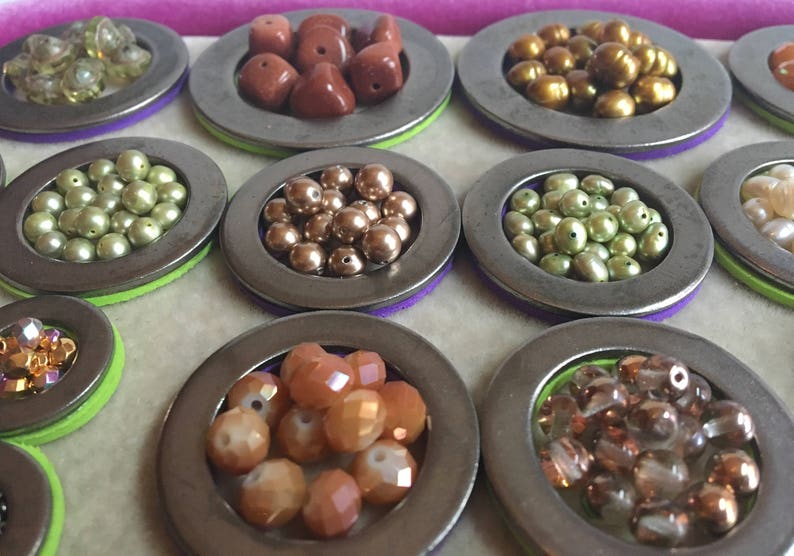 Puede incluir: Una variedad de cuentas de varios colores y formas, exhibidas en peque&ntilde;os recipientes met&aacute;licos redondos. Las cuentas incluyen tonos dorados, marrones, verdes y naranjas, algunas con apariencia facetada o perlada. Los recipientes est&aacute;n sobre fieltro morado y verde.