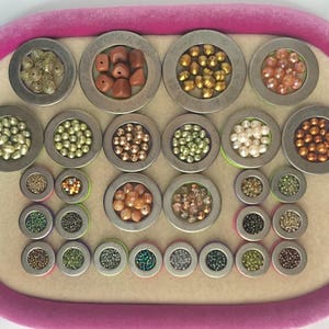 Puede incluir: Una colecci&oacute;n de cuentas de varios colores y tama&ntilde;os, exhibidas en peque&ntilde;os recipientes met&aacute;licos redondos. Las cuentas var&iacute;an de oro y cobre a verde e iridiscente, dispuestas sobre una bandeja de terciopelo rosa.