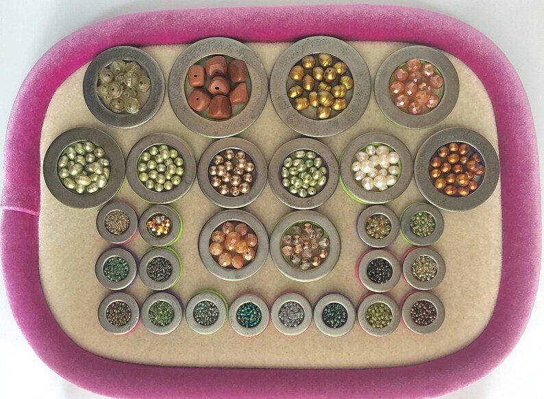Puede incluir: Una colecci&oacute;n de cuentas de varios colores y tama&ntilde;os, exhibidas en peque&ntilde;os recipientes met&aacute;licos redondos. Las cuentas var&iacute;an de oro y cobre a verde e iridiscente, dispuestas sobre una bandeja de terciopelo rosa.