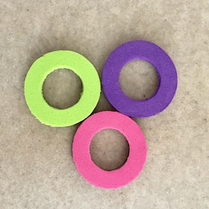 Puede incluir: Tres anillos de espuma de colores, verde lima, morado y rosa. Cada anillo tiene un agujero circular en el centro. Los anillos est&aacute;n dispuestos en un patr&oacute;n triangular sobre un fondo neutro.