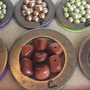 Puede incluir: Surtido de cuentas de varios colores y formas, exhibidas en peque&ntilde;os recipientes met&aacute;licos redondos. Las cuentas incluyen tonos dorados, verdes y marrones, as&iacute; como piedras de color marr&oacute;n rojizo. Cada recipiente est&aacute; etiquetado con una letra.