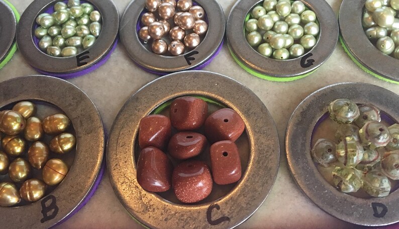 Puede incluir: Surtido de cuentas de varios colores y formas, exhibidas en peque&ntilde;os recipientes met&aacute;licos redondos. Las cuentas incluyen tonos dorados, verdes y marrones, as&iacute; como piedras de color marr&oacute;n rojizo. Cada recipiente est&aacute; etiquetado con una letra.