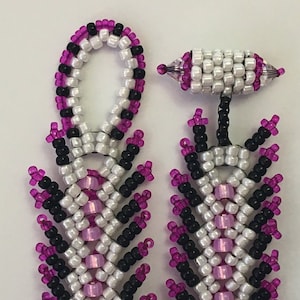 May include: Close-up of beaded earrings in a feather design. The earrings feature white, black, and pink beads. The top loop is white, black, and pink. The bottom portion has a feather-like design with the same colour scheme.