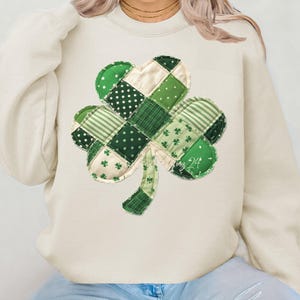 May include: Cream-colored sweatshirt featuring a large shamrock design. The shamrock is composed of various green fabric patches with polka dots, stripes, and floral patterns. The text "roz 24" is visible on the shamrock.