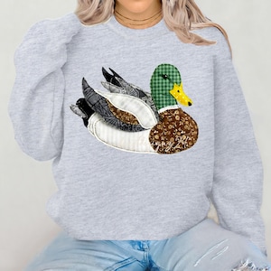 May include: A pale grey sweatshirt with a large appliqué duck design. The duck has a green plaid head, a yellow beak, and a brown floral patterned body. The wings are a mix of grey, black, and white fabrics.