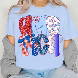 Op de afbeelding: Een lichtblauw T-shirt met het woord "America" in rode, witte en blauwe glitterletters. De letters zijn versierd met sterren, strepen en andere patriottische ontwerpen.