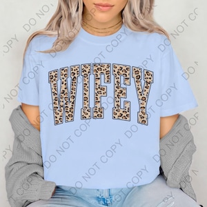 May include: Light blue t-shirt with the word "WIFEY" in large, leopard print letters. The text is outlined in a dark brown color. The shirt is short-sleeved and made of a soft material.