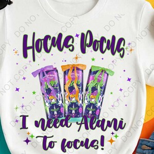 Op de afbeelding: Wit T-shirt met de tekst "Hocus Pocus" in paars en groen. Onder de tekst staan drie energiedrankblikjes met strikken. De tekst "I need Alani to focus!" staat eronder.