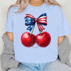 Op de afbeelding: Een lichtblauw T-shirt met een rood-wit-blauwe strik gemaakt van stof met sterren en strepen. De strik is om twee rode kersen gebonden.