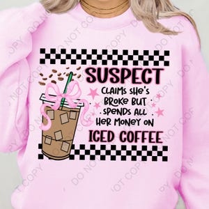 Op de afbeelding: Roze sweatshirt met een zwart-wit geruit patroon, een cartoon-ijs koffie met een roze strik en de tekst "SUSPECT CLAIMS SHE'S BROKE BUT SPENDS ALL HER MONEY ON ICED COFFEE".