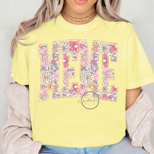 May include: A yellow t-shirt with the word "ME" in large, bold letters. The letters are filled with a floral pattern of pink, white, and orange flowers.