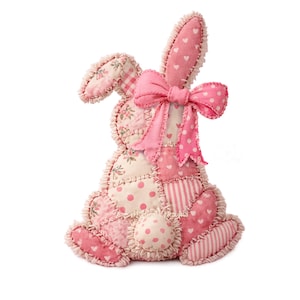 May include: A decorative patchwork Easter bunny in pink and cream. The bunny has a large pink bow, heart and polka dot patterns, and a ruffled edge. The design evokes a vintage, handmade style, perfect for Easter.