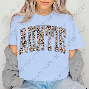 May include: Light blue t-shirt with the word "AUNTIE" in a leopard print design. The text is arched across the front of the shirt. The shirt has short sleeves and a crew neck.