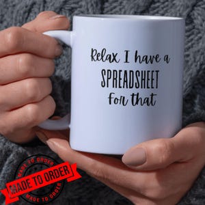Puede incluir: Una taza de cerámica blanca con el texto impreso en negro "Relax I have a SPREADSHEET for that" en una combinación de fuentes manuscritas y sans-serif. La taza tiene un acabado liso y un asa curva, presentada en una vista clara y centrada.