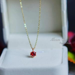 Puede incluir: Un delicado collar de cadena de oro con un colgante de piedra roja. El colgante es de forma ovalada y está engastado en un engaste de oro.