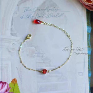 May include: A delicate gold-toned bracelet featuring a small, round, red gemstone at the center. The bracelet has a chain design with a teardrop-shaped red charm at the top. The text "Moon Cat Original design" is visible.