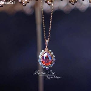 May include: A gold-colored necklace with a teardrop-shaped pendant. The pendant features a large, faceted red gemstone surrounded by small pearls and iridescent crystals. The necklace has the text "Moon Cat Original design".