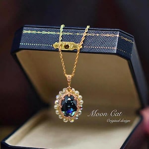 Op de afbeelding: Een goudkleurige ketting met een grote, ovale, donkerblauwe hanger. De hanger is omgeven door kleine parels en heldere kristallen. De ketting wordt gepresenteerd in een donkerblauwe sieradendoos. De tekst "Moon Cat Original design" is zichtbaar.