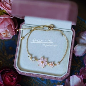 Op de afbeelding: Een goudkleurige armband met roze bloemen en heldere kristallen accenten, gepresenteerd in een roze sieradendoosje. De armband heeft de tekst "Moon Cat Original design". Het ontwerp is delicaat en vrouwelijk.