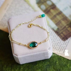 May include: A gold-colored bracelet with a large, oval-shaped emerald green gemstone at the center. The bracelet has a delicate chain design with small gold beads and a smaller green gemstone at the clasp. The bracelet is displayed on a light pink surface.