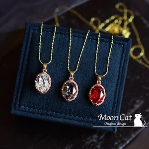 Może przedstawiać: Trzy złote naszyjniki z owalnymi zawieszkami ozdobionymi przezroczystymi, szarymi i czerwonymi kryształami. Naszyjniki są prezentowane na ciemnoniebieskim aksamitnym tle. Tekst "Moon Cat Original design" jest widoczny na dole obrazu.