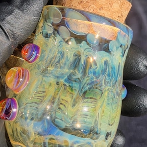 Könnte beinhalten: Ein mundgeblasenes Glasgefäß mit einem Korken. Das Gefäß weist wirbelnde Muster aus Blau, Gelb und Grün auf. Dekorative, farbenfrohe Glasperlen sind an der Seite angebracht. Das Gefäß ist etwa 7.6 cm hoch.