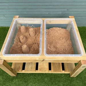 May include: A wooden sand and water table with two clear plastic bins. One bin contains brown sand with sandcastles, the other is filled with loose brown sand. The table is made of light-colored wood and sits on a green surface.