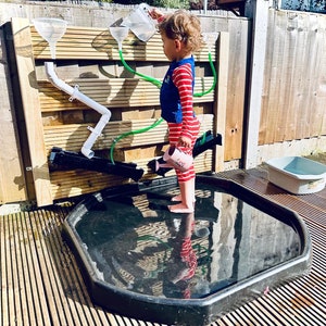 May include: A child plays with a water table, pouring water from a pitcher into a funnel. The water table is made of wood and has a variety of pipes and funnels. The child is wearing a red and white striped shirt and blue vest.