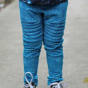 Może przedstawiać: Dziecko w niebieskich legginsach z białym wzorem kwiatowym i czarnych butach sportowych. Legginsy są zebrane przy kolanach.