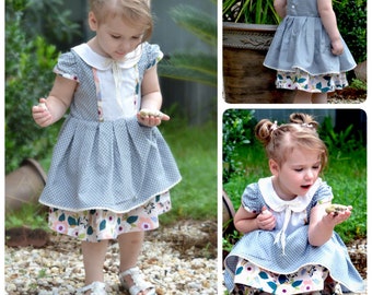Puede incluir: Una niña pequeña con un vestido de lunares azules y blancos con una falda interior de estampado floral. El vestido tiene un cuello blanco Peter Pan y mangas cortas. La niña está de pie sobre un lecho de pequeñas piedras.