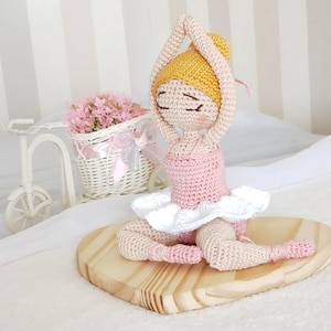 Pode incluir: Boneca bailarina de crochê em pose sentada, usando collant rosa e tutu branco. A boneca tem um coque amarelo e está sentada em uma base de madeira em forma de coração. Uma pequena bicicleta branca com uma cesta de flores rosas está ao fundo.