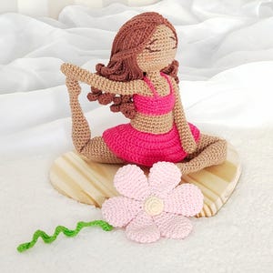 Pode incluir: Boneca de crochê feita à mão em pose de ioga, usando top e saia rosa. A boneca tem cabelo castanho e está sentada em uma base de madeira. Uma flor rosa e uma videira verde estão ao lado da boneca.