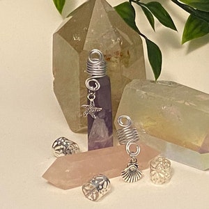 Puede incluir: Un conjunto de cuentas para el cabello plateadas con varios encantos, incluyendo una estrella de mar, una concha y un cristal morado. Las cuentas se muestran sobre una superficie blanca con cristales de cuarzo transparentes.