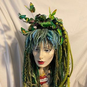 May include: A mannequin head with a striking headdress. The headdress includes green and blue dreadlocks, decorated with green and yellow butterflies. The face has red lipstick and a tattoo on the chest.