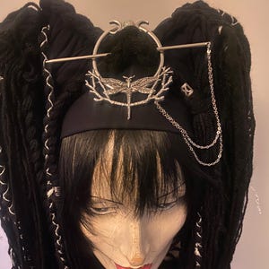 Op de afbeelding: Een zwarte pruik met lange, dikke vlechten, gestyled in twee hoge paardenstaarten. De pruik heeft een zwarte hoofdband versierd met een zilveren libel en ketting. De mannequinhoofd heeft rode lippenstift en een gedetailleerd gezicht.