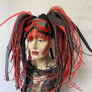 May include: A mannequin head is adorned with a black headband, red and black pigtails, and red-lensed goggles. The mannequin wears a black, distressed top with red accents. The overall aesthetic is a blend of cyberpunk and alternative fashion.