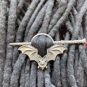 May include: Silver bat hair clip with a detailed bat design. The clip is attached to a silver bar with a textured finish. The clip is shown in a close-up view against a background of black dreadlocks.