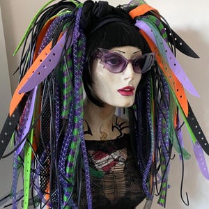 Puede incluir: Una cabeza de maniquí con cabello negro, gafas de sol moradas y extensiones de cabello vibrantes y coloridas. Las extensiones presentan detalles en negro, verde, morado y naranja. El maniquí lleva una camiseta negra con un diseño gráfico.