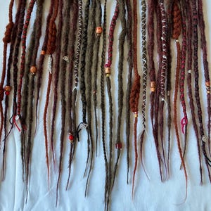 May include: An assortment of brown and reddish-brown dreadlocks, some adorned with beads and decorative accents. The dreadlocks vary in color and thickness, with some featuring intricate wrapping and embellishments. The arrangement suggests a display of handmade hair extensions.
