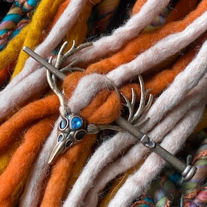 May include: A silver-toned hair accessory featuring a deer-like head with blue and black accents, set against a backdrop of colorful, textured dreadlocks in shades of orange, yellow, and pink. The accessory has a long, straight pin.