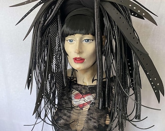 Könnte beinhalten: Eine schwarze Perücke mit langen, schwarzen, geflochtenen Extensions. Die Extensions bestehen aus einem Material, das Leder ähnelt. Die Perücke ist zu zwei hohen Pferdeschwänzen gestylt. Die Perücke wird von einer Mannequin mit einem schwarzen Netz-Oberteil getragen.