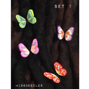 Op de afbeelding: Set van vier kleurrijke vlinder haarclips. De vlinders zijn groen, roze, paars en rood met zwarte contouren. De haarclips bevinden zich op een zwarte achtergrond. De tekst "SET 1" en "MISSSNEEDLES" is zichtbaar in de afbeelding.